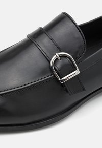 Černá kožená mokasína se stříbrnou kovovou sponou a viditelným šitím podél špičky a boků.