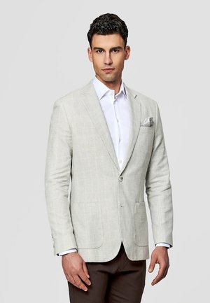 Mężczyzna w jasnoszarej kratkowanej marynarce, białej koszuli wizytowej i ciemnobrązowych spodniach, stojący na tle gładkiego szarego tła.