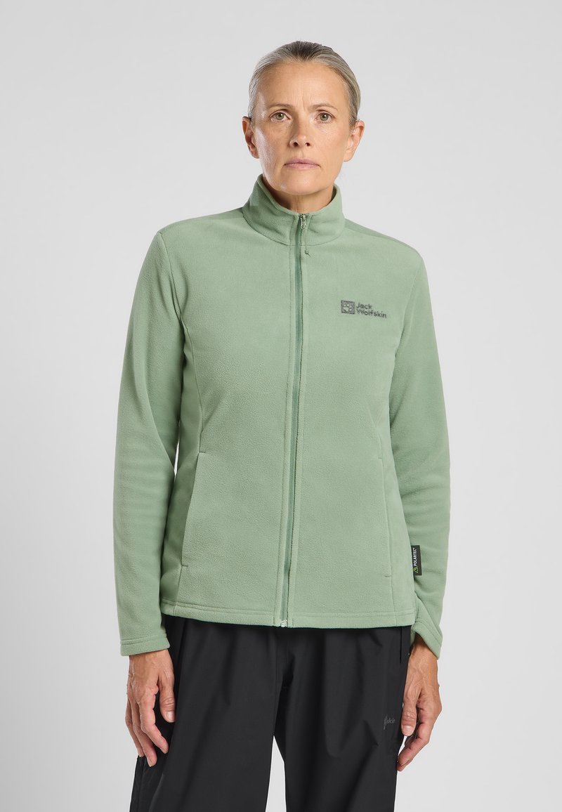 Green fleece jacket with a high collar, front zipper, and a logo on the chest. Smooth texture and fitted design, paired with black pants.