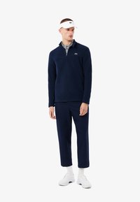 Pullover navy in morbido materiale con colletto a mezza zip, caratterizzato da un colletto interno a righe e logo Lacoste. Abbinato a pantaloni navy coordinati e scarpe bianche.