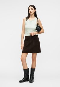 Frau trägt ein hellgrünes ärmelloses Oberteil, einen dunkelbraunen Rock mit Taschen, schwarze Stiefel bis zur Mitte der Wade und eine kleine schwarze runde Handtasche.