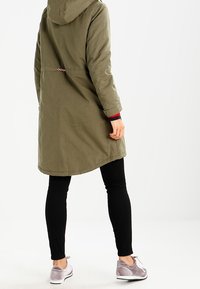 Parka verde oliva con capucha, que cuenta con cintura ajustable y bolsillos laterales. Combinada con leggings negros y zapatillas deportivas grises.