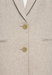 Beige herringbone-patterned blazer with notched lapels, two gold-tone buttons, and side pockets. Textured fabric, tailored fit.