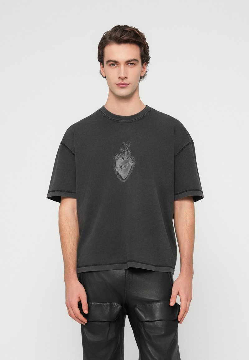 Μαύρο φαρδύ t-shirt από βαμβάκι, με μπροστινό σχέδιο καρδιάς και λεπτομέρειες φυλλώματος. Κοντά μανίκια, στρογγυλή λαιμόκοψη και ακατέργαστο τελείωμα.