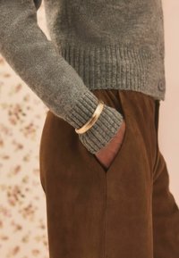 Grå strikket sweater med ribbede manchetter, parret med brune ruskindsbukser; inkluderer et lagdelt armbånd i guldtonet på håndleddet.