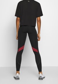 Černé sportovní legíny s kontrastními červenými diagonálními pruhy, prošitými detaily, v kombinaci s černým krátkým tričkem a bílými teniskami s vzorem.