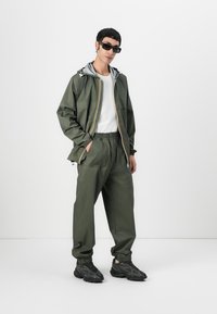 Giacca impermeabile verde oliva con cappuccio grigio e polsini elasticizzati, abbinata a pantaloni coordinati. Sneakers nere con suole testurizzate.