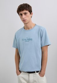 Joven con cabello corto castaño que lleva una camiseta azul claro de "The Scalpers Company" y pantalones blancos, con las manos en los bolsillos, fondo gris neutro.