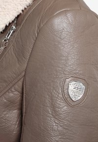 Veste en cuir marron avec une finition texturée, dotée d'un emblème en argent sur l'épaule et d'un col en fourrure de mouton.