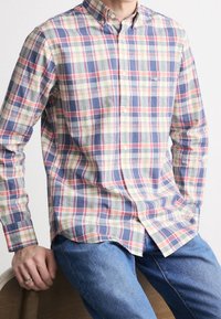 Man draagt een geruit overhemd met lange mouwen in blauw, groen en rood, gecombineerd met een blauwe spijkerbroek, zittend op een houten stoel.