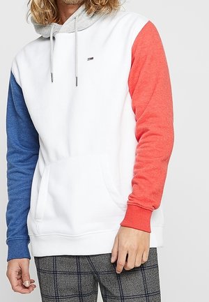 Osoba ubrana w biały kaptur z szarym kapturem, niebieskim lewym rękawem, czerwonym prawym rękawem oraz w kratkowane spodnie, stojąca na jednolitym tle.