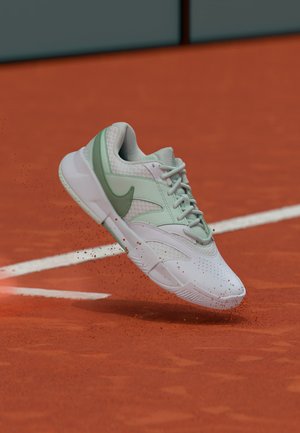 Baskets blanches et vert clair soulevant de la poussière d'argile rouge sur un court de tennis près de la ligne de fond.