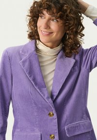 Violetti corduroy-takki, jossa on teksturoituja raitoja, lovitetut läpät, kaksi etutaskua ja kultasävytteiset napit; kerroksittain vaalean turtlenechin päällä.