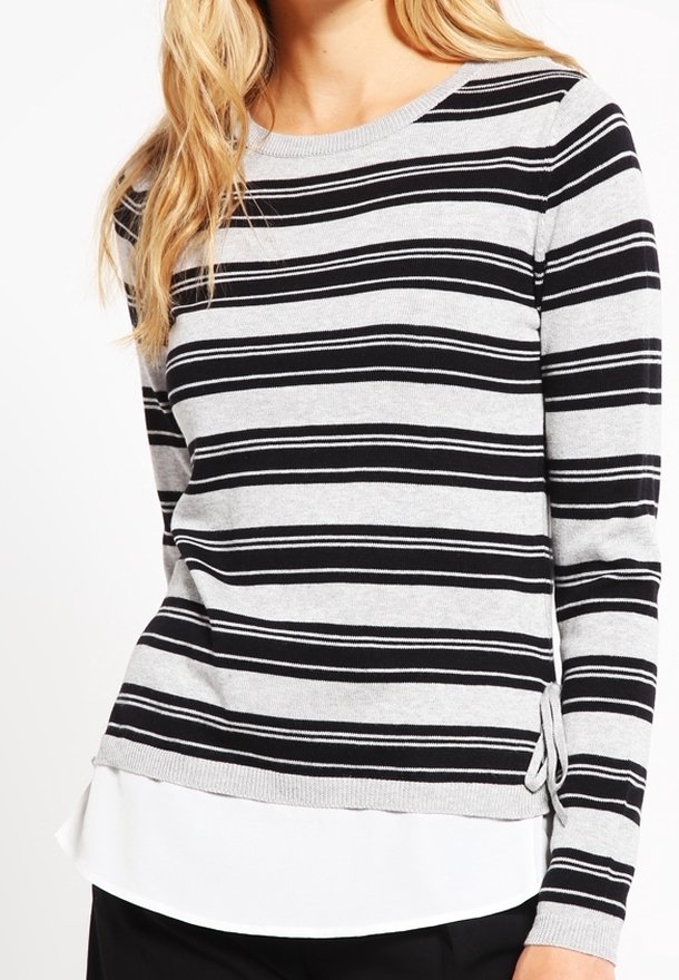 Femme portant un pull à manches longues rayé horizontalement gris et noir par-dessus un chemisier blanc à ourlet superposé.