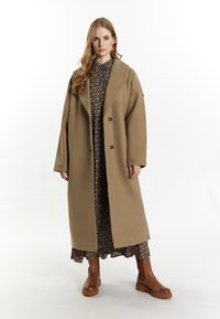 Beige kabát z vlněné směsi s velkým límcem, dvěma knoflíky a širokými rukávy. Nosený přes květinové šaty; hnědé kotníkové boty.