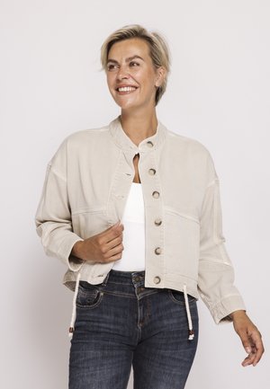 Lächelnde Frau trägt eine hellbeige, geknöpfte Jacke über einem weißen Top und dunkelblauen Jeans vor einem einfarbigen hellen Hintergrund.