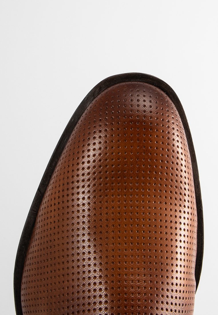 Zapato de cuero marrón con punta redonda, que presenta un diseño perforado y una textura suave. Biselado negro a lo largo de la suela.
