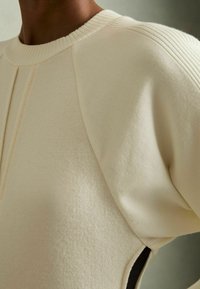 Krém színű, puha anyagból készült pulóver, bordázott gallérral és vállvarrással, kivágott részletekkel és textúrált díszítésekkel.