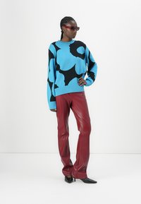 Plavi i crni uzorčasti oversized pulover, crvene kožne hlače i crne špicaste štikle; model nosi sunčane naočale, stoji na bijelom pozadini.