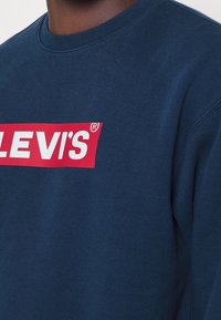 Sweat-shirt bleu marine en coton, avec un logo rectangulaire rouge audacieux portant le texte "LEVI'S®" en blanc sur le devant. Texture douce.