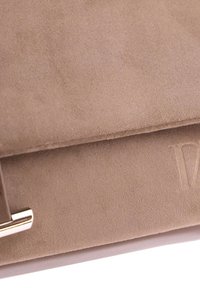 Acercamiento de la solapa de un bolso de ante beige con logo en relieve y detalle de cierre metálico dorado.