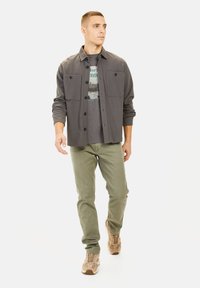 Grijze button-up shirt over een graphic t-shirt, gecombineerd met lichtgroene broek en beige sneakers. Casual, relaxed fit.