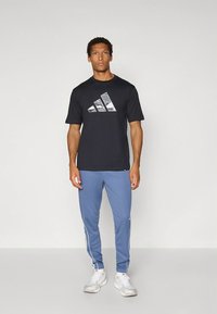 T-shirt preta de mangas curtas com logótipo reflexivo da Adidas; combinada com calças azul claro com riscas brancas nas laterais. Modelo em pose de pé.