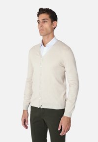 Cardigan beige con scollo a V, dotato di una chiusura con bottoni sul davanti, maniche lunghe e orlo a costine. Indossato sopra una camicia bianca e pantaloni verdi.