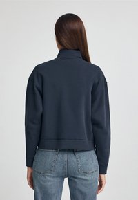 Donna con lunghi capelli castani, che indossa un felpa blu scuro e jeans blu, vista di spalle su uno sfondo chiaro e semplice.