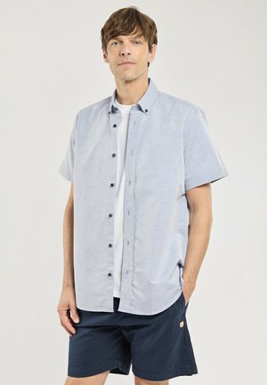 Homme portant une chemise bleu clair à manches courtes boutonnée par-dessus un T-shirt blanc et un short bleu foncé, debout avec une main dans la poche devant un fond uni.