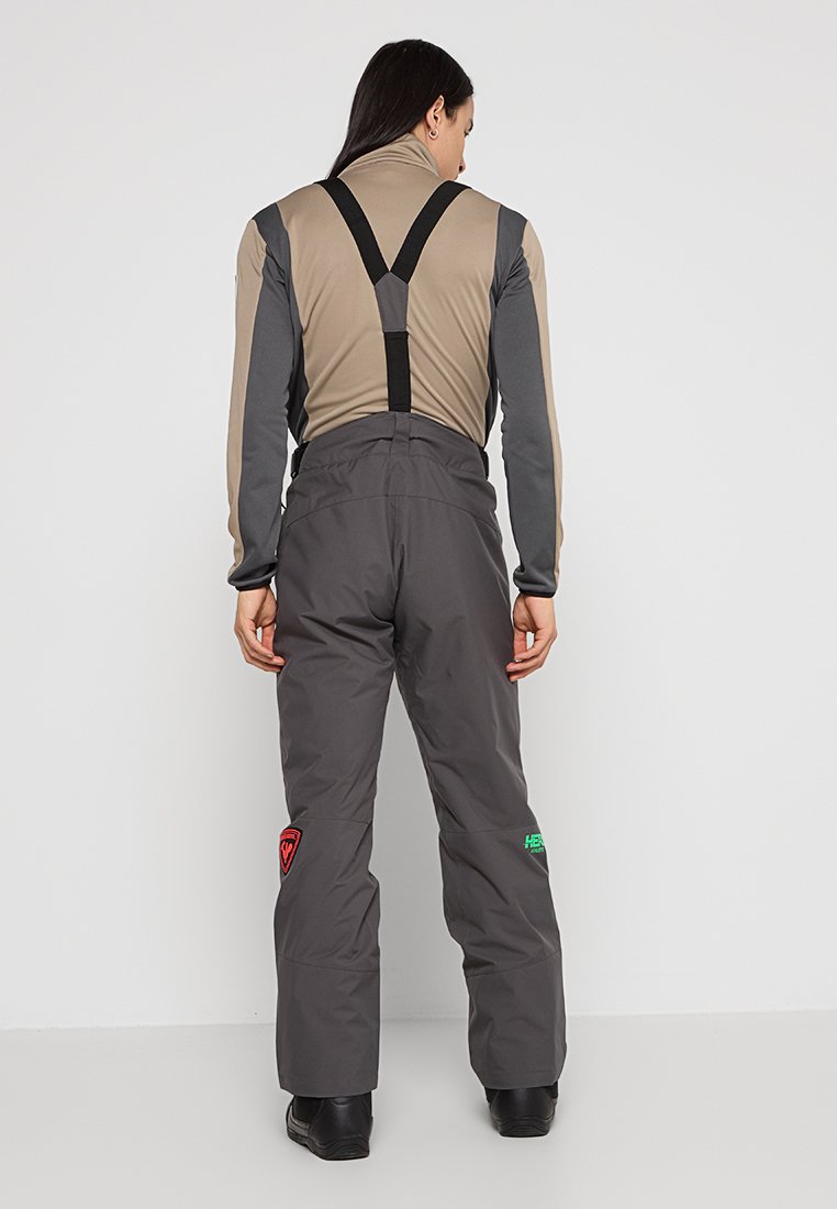 Szare spodnie śniegowe z regulowanymi czarnymi szelkami, z czerwonymi i zielonymi logo. Wykonane z trwałej tkaniny, zaprojektowane do aktywności zimowych.