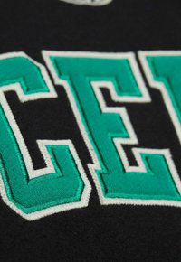 Tela negra con letras bordadas en verde y blanco de gran tamaño. La textura es suave con detalles de costura elevados alrededor de las letras.