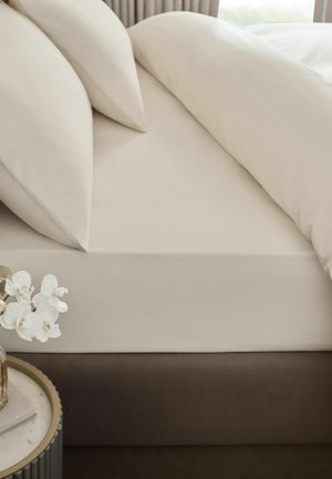 Beiges Bett mit Spannbettlaken, zwei Kissen und Bettdecke, neben rundem Tisch mit weißen Orchideen in Glasvase.
