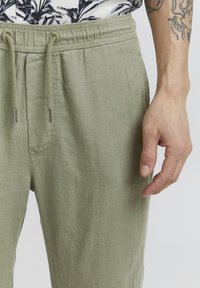 Pantalon en lin vert clair avec une taille élastique, cordon de serrage et poches latérales ; la texture semble douce et respirante.