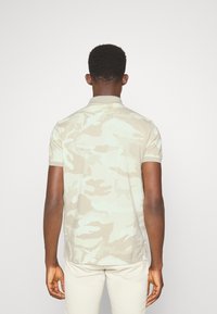 Beige kamuflážová polo košile s krátkým rukávem, límečkem a jemným texturem. Obsahuje decentní zelené akcenty a postranní rozparky.