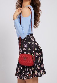 Czerwona pikowana torebka z trójkątnym logo, wyposażona w długi pasek. Noszona z niebieską bluzką i czarną spódnicą w kwiatowy wzór.