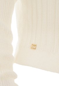 Weißer Strickpullover mit strukturiertem Design, gerippten Bündchen und einem goldenen Markenlabel, das am unteren Saum angenäht ist.