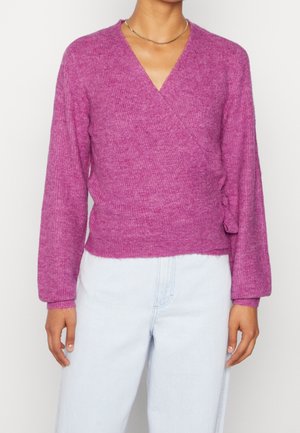 Kvinde iført en langærmet, omslagsstil, struktureret lilla sweater kombineret med lyseblå højtaljede jeans og en tynd guldkæde.