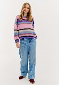Pletený svetr s růžovými, fialovými a černými pruhy, zkombinovaný s volnými světlými modrými džíny a vínovými botami. Měkká textura a uvolněný střih.