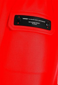 Tela roja con una textura suave, que presenta una etiqueta de parche negra con texto blanco y dos botones negros. El diseño incluye un bolsillo insertado.