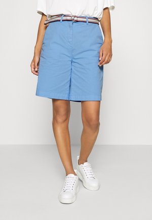 Person iført lyseblå knælange shorts med et stribet bælte, hvid t-shirt og hvide platformsneakers, stående på en grå gulv.