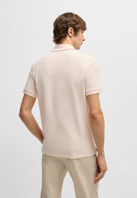 Ανοιχτό ροζ πόλο μπλουζάκι, κοντά μανίκια, λεία υφή, γιακάς και λεπτομέρειες στο τελείωμα. Φορώντας με ανοιχτό μπεζ παντελόνι. Πίσω θέα, μινιμαλιστικός σχεδιασμός.