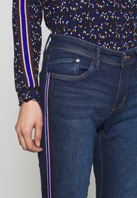 Džíny z džínoviny v tmavě modré barvě s bočními pruhy v oranžové, bílé a modré. Mají pět kapes a přiléhavý střih. Detaily s cikcakovým lemováním na rukávech.