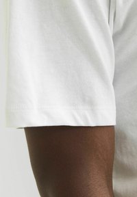 Hvid bomulds t-shirt med korte ærmer, der har en færdigkant og en glat tekstur. Den synlige arm har en mørk hudtone.