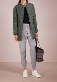 Zelená prošívaná péřová bunda s vysokým límcem, kombinovaná s šedými tepláky a bílými teniskami. Černý top a kostkovaná taška dotvářejí vzhled.