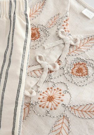 Vyšívaná tkanina so vzormi kvetov a listov v oranžovej a čiernej farbe na béžovom pozadí, doplnená o pruhovaný materiál v bielej farbe s nádychom smotany.