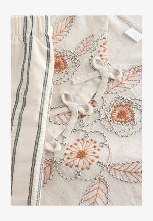 Vyšívaná tkanina so vzormi kvetov a listov v oranžovej a čiernej farbe na béžovom pozadí, doplnená o pruhovaný materiál v bielej farbe s nádychom smotany.