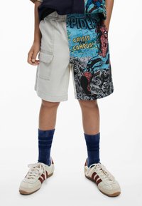 Kind met ongelijke shorts, de ene is beige in cargo-stijl en de andere heeft een Spider-Man stripprint, gecombineerd met blauwe sokken en witte sneakers.