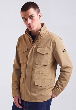 Junger Mann trägt eine hellbraune Jacke mit vier Taschen vorne und einem Aufnäher am linken Ärmel, steht vor einem schlichten hellen Hintergrund.