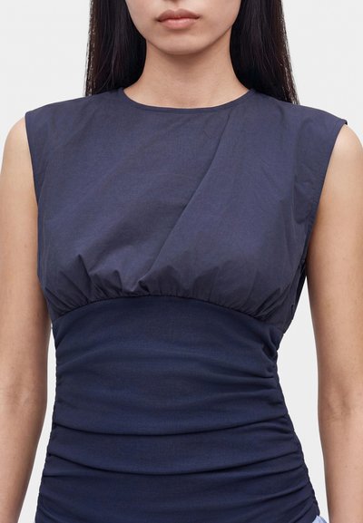 Femme portant une robe sans manches bleu marine avec un tissu froncé au niveau de la poitrine et des détails froncés autour de la taille sur un fond uni.